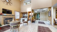 This single-floor community space combines functionality with a warm, residential aesthetic. The centerpiece of the space is a grand lounge area distinguished by dramatically soaring two-story ceilings and rich, golden-amber walls that lend the space a welcoming, sun-kissed ambiance. Light tile flooring with wood-like detailing flows throughout, unifying the various areas seamlessly.

The lounge is anchored by a classic fireplace framed in mosaic tile accents, topped with a mounted flat-screen television, and flanked by large windows with soft draping curtains that allow natural light to filter in. Comfortable seating arrangements, including cushioned chairs and accent benches, invite relaxation and conversation throughout the space.

Adjacent to the lounge, a dedicated business center features a built-in desk station set against a striking mosaic tile backsplash, complemented by upper cabinetry and framed artwork. This workstation area is thoughtfully designed for productivity while maintaining the overall aesthetic cohesion of the space.

A wide, well-lit corridor extends toward additional rooms and private spaces, lined with patterned area rugs and tasteful wall art. The main entrance features a set of rich wood double doors flanked by transom windows, reinforcing the grandeur of the double-height entry. Overall, the space exudes a professional yet approachable character, ideal for community gatherings or resident services.