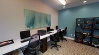 This single-floor business center is designed to offer a productive and professional environment for residents or guests. The space is anchored by a continuous white desk surface running along the wall, providing ample workspace for multiple users simultaneously. Each workstation is equipped with a desktop computer and ergonomic black office chair, ensuring comfort during extended use. A shared laser printer is conveniently positioned along the same desk surface, making document printing easily accessible to all users.

The room's interior design strikes a balance between function and aesthetics. A calming teal accent wall adds a pop of color and visual interest, complementing the neutral beige tones of the surrounding walls. A large, eye-catching abstract canvas artwork in shades of green and teal hangs prominently above the workstations, contributing to the space's modern and polished character. The patterned carpet in warm orange and cream tones adds texture underfoot and ties the overall color palette together. Overhead, a combination of flush and linear lighting ensures the workspace is well-lit throughout. A bookshelf along one wall provides additional utility. Overall, the space is thoughtfully arranged to support focused, efficient work in a clean and contemporary setting.