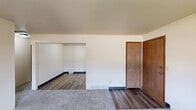This single-floor residential unit offers a practical and well-organized layout designed for comfortable everyday living. The galley-style kitchen is a standout feature, lined with warm wood-toned cabinetry on both sides providing generous storage space. It is equipped with a white electric range, refrigerator, and a double sink set into light-colored countertops, all finished with light wood-style flooring that extends through the entry and adjoining areas.

The living area is generously sized, featuring soft gray carpet underfoot and a window that allows natural light to filter in while offering views of the surrounding neighborhood. A wall-mounted HVAC unit provides climate control. The space flows naturally into additional carpeted rooms, including multiple bedrooms, each offering good proportions, neutral wall tones, and access to closet storage — one of which features a walk-in style closet.

The full bathroom is well-appointed with a wood-cabinet vanity, a wide mirror with overhead lighting, a combination tub and shower, and matching wood-finish built-in shelving. Consistent wood laminate flooring ties the bathroom together with the rest of the unit's non-carpeted zones.

Overall, the unit presents a clean, neutral palette with consistent warm wood accents, offering a functional and move-in ready living environment.