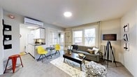 This single-floor space is thoughtfully arranged to maximize functionality within a compact footprint. The main living area serves as the centerpiece, featuring a charcoal gray sofa paired with a patterned area rug and a dark wood coffee table, creating a grounded, modern aesthetic. Two vibrant yellow chairs and a small round bistro table occupy a corner near the kitchenette, offering a casual dining or lounging nook that adds a pop of color to the neutral palette.

Large windows along the exterior wall flood the living space with natural light, offering views of outdoor greenery and contributing to an open, airy feel. A wall-mounted mini-split HVAC unit ensures year-round climate control. Overhead, a flush-mount ceiling light provides clean, even illumination.

The kitchenette, positioned along one wall, includes white cabinetry, a dark countertop, a microwave, and a compact sink — efficiently designed for everyday use. French glass-paned doors lead to a separate bedroom, providing privacy while maintaining visual connectivity and light flow between spaces. The overall ambiance is contemporary and functional, with a neutral gray and white base accented by warm, bold furnishing choices.