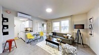 This single-floor space is thoughtfully arranged to maximize functionality within a compact footprint. The main living area serves as the centerpiece, featuring a charcoal gray sofa paired with a patterned area rug and a dark wood coffee table, creating a grounded, modern aesthetic. Two vibrant yellow chairs and a small round bistro table occupy a corner near the kitchenette, offering a casual dining or lounging nook that adds a pop of color to the neutral palette.

Large windows along the exterior wall flood the living space with natural light, offering views of outdoor greenery and contributing to an open, airy feel. A wall-mounted mini-split HVAC unit ensures year-round climate control. Overhead, a flush-mount ceiling light provides clean, even illumination.

The kitchenette, positioned along one wall, includes white cabinetry, a dark countertop, a microwave, and a compact sink — efficiently designed for everyday use. French glass-paned doors lead to a separate bedroom, providing privacy while maintaining visual connectivity and light flow between spaces. The overall ambiance is contemporary and functional, with a neutral gray and white base accented by warm, bold furnishing choices.