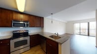 This single-floor residence is designed with an open-concept layout that connects the kitchen and living area seamlessly. The kitchen is well-equipped with rich dark wood cabinetry, granite countertops, a white subway tile backsplash, and a full suite of stainless steel appliances, including a refrigerator, electric range, built-in microwave, and dishwasher. A peninsula with an integrated sink and pendant lighting above serves as both a functional workspace and a natural divider between the kitchen and the adjoining living area. The living area features warm hardwood-style flooring and neutral walls, creating a bright and inviting atmosphere. A sliding glass door opens directly onto a private balcony, allowing natural light to fill the space and providing views of the surrounding area. The bedroom is carpeted for comfort and includes large windows and a sliding door to an additional outdoor space, further enhancing natural light and ventilation. The bathroom is finished with tile flooring and features a full soaking tub with a tiled surround, complemented by granite-topped vanity cabinetry. The overall design maintains a clean, cohesive aesthetic throughout.