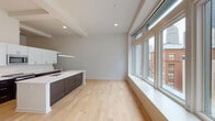 This single-floor urban loft masterfully combines industrial heritage with contemporary design. Light hardwood flooring flows throughout the open-plan living and kitchen area, anchored by a sleek kitchen island with dark cabinetry and white quartz countertops. The kitchen is fully equipped with stainless steel appliances, including a refrigerator, built-in microwave, and range, complemented by crisp white upper cabinets and ample storage. A striking exposed brick wall and visible ductwork add raw, industrial character that contrasts beautifully with the clean, modern palette. Large windows along the main living area flood the space with natural light and frame city views. The bedroom features soaring ceilings, plush carpeting, and a dramatic arched window that draws in additional natural light and urban scenery. A stepped entryway connects the bedroom to the rest of the space, adding architectural interest. Two bathrooms are finished with dark tile flooring, floating dark-wood vanities, and white countertops. One bathroom includes a bathtub with tiled surround, while the other offers tall windows overlooking the cityscape. A ceiling fan provides comfortable airflow throughout the living space, and recessed lighting ensures a well-lit environment throughout.