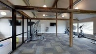 This single-story fitness center combines rustic architectural charm with functional modern amenities. The space is defined by its striking exposed wooden timber beams and posts, which frame the vaulted ceiling and lend a warm, barn-inspired aesthetic to the interior. Visible ductwork runs along the ceiling, adding an industrial contrast to the natural wood elements, while pendant globe lights provide consistent, even illumination throughout the space.

The flooring consists of modular carpet tiles arranged in a varied pattern of blues, grays, and neutral tones, offering comfort underfoot during workouts. The facility is equipped with a multi-station cable weight machine, stationary cardio equipment including an upright bike and elliptical trainer, and a wall-mounted flat-screen television for entertainment during exercise sessions. A water fountain station is conveniently positioned along the back wall.

Large glass sliding doors on one side allow natural light to filter into the space, while the white walls and high ceiling contribute to an open, airy feel. The overall ambiance balances rustic character with a clean, practical fitness environment.