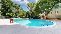 Located at 3640 16th St NW in Washington, DC, this outdoor amenity space centers around a generously sized swimming pool with a curved, freeform design and crystal-clear blue water. The pool is surrounded by a broad concrete sundeck that provides ample room for lounging, with seating options including reclining lounge chairs, plastic patio chairs, and umbrella-shaded tables for relaxed outdoor gatherings. The area is framed by a multi-story light brick residential building, whose many windows overlook the pool courtyard, creating an enclosed and private atmosphere. Mature trees line the perimeter of the space, offering natural shade and a lush, park-like setting that softens the urban environment. A dedicated pool house structure along the building's exterior provides access to pool equipment and storage. An additional patio courtyard adjacent to the pool offers further open-air seating and gathering space, enclosed by the building's brick facade and accented with potted plants. Iron railings and fencing define the boundaries of the deck area, while the abundant greenery and open sky above create a refreshing retreat in the heart of Washington, DC's 16th Street corridor.