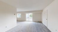 This single-floor residence offers a straightforward and functional layout organized around a generously sized main living area. The living room is a standout feature, with plush grey carpeting underfoot, neutral cream-toned walls, and multiple windows that invite abundant natural light. A sliding glass door opens directly onto a private balcony with a white railing, offering outdoor views of surrounding greenery. A side window further enhances the brightness of the space.

The kitchen is positioned adjacent to the living area and is defined by dark espresso-toned cabinetry contrasted against light countertops. White appliances, including a refrigerator, electric range, and dishwasher, are neatly arranged alongside an under-mount sink. Light wood-tone vinyl flooring distinguishes the kitchen from the carpeted areas throughout the rest of the unit.

The bedroom offers a comfortable footprint with soft grey carpet, a standard closet with bifold doors, and a window providing natural daylight and outdoor views. An additional carpeted room with a window provides flexible use as a second bedroom or office space.

The bathroom is clean and well-appointed, featuring a white vanity with brushed nickel hardware, a toilet, and a full tub-shower combination with a light vinyl floor.