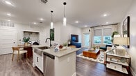 This well-designed single-floor residence showcases a thoughtful open-concept layout that seamlessly connects the kitchen, dining, and living areas. The kitchen is a standout feature, equipped with white shaker cabinetry accented by matte black hardware, quartz countertops, stainless steel appliances including a dishwasher and range, subway tile backsplash, a central island with a farmhouse-style sink, and stylish pendant lighting overhead. A dedicated dining area sits adjacent to the kitchen, with a convenient in-unit laundry closet nearby. The living room is bright and inviting, benefiting from large windows and a glass door that flood the space with natural light and provide outdoor views. Dark hardwood-style flooring runs consistently throughout, grounding the neutral color palette. The primary bedroom is generously sized with large windows, ample natural light, and direct access to an en-suite bathroom featuring a sliding glass shower enclosure, white vanity, and a walk-in closet. A second bedroom offers similar brightness and comfort. The second bathroom is equally refined, with a backlit vanity mirror, white cabinetry, and a tub-shower combination. Together, the spaces deliver a cohesive, modern aesthetic with high-quality finishes at every turn.