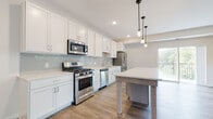 This single-floor residence combines modern finishes with a functional layout. The kitchen is a standout feature, showcasing white shaker cabinetry, light quartz countertops, and a soft blue-grey subway tile backsplash. A full suite of stainless steel appliances—including a gas range, built-in microwave, dishwasher, and French door refrigerator—lines the main wall, while a freestanding island provides additional prep and dining space. Pendant lights overhead add a warm, stylish accent. The open living area features wood-look plank flooring and multiple windows that invite natural light and outdoor views of surrounding greenery. The bedroom is finished with plush carpet and a bold blue accent wall that adds character to the space. A walk-in closet with wire shelving provides generous storage. Two bathrooms serve the unit: one featuring a glass-enclosed walk-in shower with marble-style tile surround and wood-look flooring, paired with a taupe vanity; the other offering a clean white soaking tub with matching surround. Both bathrooms maintain a cohesive, polished aesthetic that complements the rest of the home's contemporary design.