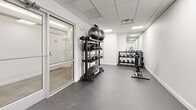 Located at 10 Dorrance Street in the heart of Providence, Rhode Island, this fitness center offers a well-appointed and thoughtfully organized workout facility across a single floor. The space features a clean, modern aesthetic defined by bright white walls, a dropped ceiling with recessed panel lighting, and durable dark rubber flooring throughout — creating a functional yet polished environment suited for a range of fitness routines.

The cardio area is equipped with multiple machines, including a treadmill, elliptical trainer, and stationary cycle, each outfitted with individual digital display screens. This section is separated from an adjoining space by a full-height glass partition with a sliding glass door, allowing visual openness while maintaining distinct zones within the facility.

The free weights area is positioned toward the rear of the space and features a multi-tiered dumbbell rack stocked with a variety of weights, an adjustable workout bench, and a full-length mirror that visually expands the room. An additional functional training zone includes a tall rack holding kettlebells, medicine balls, and balance equipment, catering to dynamic and strength-based workouts.

The overall ambiance is bright, clean, and minimalist, with ample overhead lighting ensuring a well-illuminated environment throughout. The layout maximizes usability while keeping the space uncluttered and accessible.