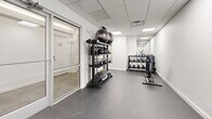 Located at 10 Dorrance Street in the heart of Providence, Rhode Island, this fitness center offers a well-appointed and thoughtfully organized workout facility across a single floor. The space features a clean, modern aesthetic defined by bright white walls, a dropped ceiling with recessed panel lighting, and durable dark rubber flooring throughout — creating a functional yet polished environment suited for a range of fitness routines.

The cardio area is equipped with multiple machines, including a treadmill, elliptical trainer, and stationary cycle, each outfitted with individual digital display screens. This section is separated from an adjoining space by a full-height glass partition with a sliding glass door, allowing visual openness while maintaining distinct zones within the facility.

The free weights area is positioned toward the rear of the space and features a multi-tiered dumbbell rack stocked with a variety of weights, an adjustable workout bench, and a full-length mirror that visually expands the room. An additional functional training zone includes a tall rack holding kettlebells, medicine balls, and balance equipment, catering to dynamic and strength-based workouts.

The overall ambiance is bright, clean, and minimalist, with ample overhead lighting ensuring a well-illuminated environment throughout. The layout maximizes usability while keeping the space uncluttered and accessible.