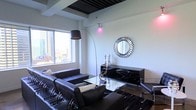 This sophisticated single-floor space showcases a refined contemporary aesthetic defined by a cohesive dark color palette and high-contrast design elements. The living area serves as the centerpiece, anchored by a set of rich black leather seating arranged around a glass-top coffee table, creating an atmosphere of polished elegance. A dramatic arc floor lamp with a large black shade curves gracefully over the seating area, complementing the recessed ceiling detail above, which features a dark inset panel that adds architectural depth. A decorative sunburst mirror with a metallic silver frame adorns the wall, serving as a striking focal point alongside a sleek, high-gloss black media console accented with chrome hardware. Sculptural silver décor pieces add refined detail to the space. A large window floods the room with natural light and frames a sweeping urban skyline view, reinforcing the elevated, city-living character of the space. The overall ambiance is bold, curated, and contemporary — ideal for those who appreciate modern design with an upscale, metropolitan edge.