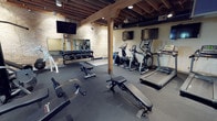 This fitness center combines rustic architectural character with a full suite of modern exercise equipment. The space is defined by its exposed wooden beam ceiling structure and warm wood posts, which lend an industrial-loft aesthetic to the environment. Brick accent walls add texture and visual interest throughout, while rubber flooring throughout the workout areas provides safety and durability underfoot.

The cardio section is well-appointed with multiple treadmills, elliptical trainers, and stationary bikes, each positioned for comfortable use with wall-mounted flat-screen televisions nearby for entertainment. The strength training area features a variety of cable machines and weight machines, as well as adjustable benches and a fully stocked dumbbell rack with a large wall mirror, allowing users to monitor form during workouts. Stability balls are also available for functional training exercises.

Natural light filters into the space through a row of windows along one wall, brightening the strength equipment area. An entry area near the fitness center's access point includes a water station and informational signage, providing a welcoming transition into the workout space. The overall ambiance is functional yet characterful, blending the warmth of exposed timber construction with the practicality of a comprehensively equipped fitness facility.