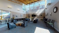 This single-story community space is defined by its striking architectural design, most notably its soaring vaulted ceilings supported by exposed wooden beams and a dramatic wall of clerestory windows that flood the interior with abundant natural light. The patterned blue and gold carpet anchors the main lounge area, which is thoughtfully arranged with multiple seating groupings — including plush blue sofas, accent chairs, and a distinctive glass-topped table with a natural wood base — encouraging both social interaction and quiet relaxation. Freestanding shelving units with a sleek metallic finish serve as room dividers, adding visual interest while maintaining an open, airy feel. A built-in bookcase along one wall provides a dedicated reading nook, complemented by a wall-mounted flat-screen television nearby. A gallery of framed photographs and artwork is displayed throughout, adding a curated, community-oriented character to the space. A casual kitchenette area features a pass-through counter with bar seating and is equipped with a popcorn machine, lending a welcoming, social atmosphere. Additional amenities include computer workstations positioned near a bright window, as well as a quieter seating alcove ideal for conversation or media viewing. The overall ambiance is warm, modern, and inviting.