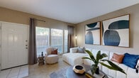 This single-floor residence offers a cohesive and stylishly appointed living experience. The entryway opens into a welcoming living room bathed in natural light from large windows dressed with elegant draping curtains. The space is anchored by plush carpeting and a white modular sofa, complemented by curated abstract wall art in gold-framed panels. A white brick fireplace serves as a striking focal point, adding warmth and character to the room.

Adjacent to the living area, the dining space features a round marble-top table surrounded by sleek dark chairs, accented by bold, colorful framed artwork on neutral-toned walls. The kitchen is outfitted with white cabinetry, light stone-effect countertops, and hardwood-style flooring, providing ample storage and counter space for everyday living.

The bedroom is generously proportioned, featuring soft carpeting, a ceiling fan, rich jewel-toned drapery, and vibrant large-scale artwork that adds an energetic focal point to the serene retreat. The bathroom includes an extended vanity with white cabinetry, a wide mirror with overhead lighting, and clean, bright finishes throughout.

Together, these spaces create a comfortable and contemporary home with a warm, curated aesthetic.