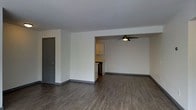 This single-floor space presents a clean, modern aesthetic defined by dark hardwood-style flooring that runs throughout the open living area, creating a sleek and cohesive look underfoot. The neutral white walls and light-toned ceiling provide a bright, airy backdrop that enhances the sense of openness across the layout. A stylish ceiling fan with an integrated light fixture is centrally positioned in the main living area, offering both comfort and illumination. The space flows naturally into an adjoining area, visible through an open pass-through or partial wall, where upper cabinetry hints at a kitchen space equipped with ample storage. Dark gray baseboards provide a subtle but defined contrast against the lighter walls, adding a contemporary design touch throughout. A gray interior door on the far side of the room contributes to the cohesive color palette while offering access to an additional area of the space. The overall layout is open and versatile, providing an inviting canvas well-suited for a variety of furniture arrangements and functional uses. The combination of hard flooring, neutral tones, and modern fixtures gives this space a fresh, move-in-ready character.