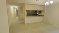 This two-floor residential space offers a functional and open layout designed for comfortable everyday living. The main living area is finished with neutral wall-to-wall carpeting that extends throughout, creating a cohesive and understated aesthetic. A defining feature of the space is the pass-through kitchen, which opens directly into the living area. The kitchen is equipped with stainless steel appliances, including a refrigerator and microwave, complemented by warm wood-toned cabinetry and dark countertops that provide a subtle contrast to the light-colored walls and flooring. The pass-through counter extends slightly into the living space, offering a casual surface for dining or entertaining. A long hallway connects the living area to additional rooms, suggesting a well-organized floor plan with clear separation between living and private spaces. Recessed and ceiling-mounted lighting provides consistent illumination throughout. The neutral color palette of cream and beige tones contributes to a calm, move-in-ready ambiance. The second floor adds additional living space, making this a versatile and practical multi-level home suited for a range of lifestyles.