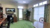 This single-story space exudes a refined, professional character through its thoughtfully curated design elements. The office is finished with warm, cream-toned walls accented by crisp crown molding, lending a sense of traditional elegance throughout. Lush green carpet covers the floor, adding warmth and a touch of color that complements the overall palette of the room.

Natural light flows generously through multiple windows dressed with patterned, floor-length drapery panels and white wood blinds, creating a bright yet comfortable environment. Transom windows above the main windows further enhance the sense of openness and allow additional light to fill the space.

The office is thoughtfully furnished with a classic executive desk and leather chair, accompanied by a dark wood chest of drawers topped with a traditional table lamp. A large, framed landscape painting adorns one wall, adding a sophisticated artistic focal point. A tall, lush indoor palm plant positioned in the corner introduces a natural, vibrant element to the interior. Overhead, a flush-mounted skylight panel supplements the natural light from the windows, ensuring the space remains consistently well-lit. Together, these details create a polished, inviting office environment suited for focused, professional work.