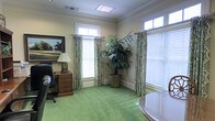 This single-story space exudes a refined, professional character through its thoughtfully curated design elements. The office is finished with warm, cream-toned walls accented by crisp crown molding, lending a sense of traditional elegance throughout. Lush green carpet covers the floor, adding warmth and a touch of color that complements the overall palette of the room.

Natural light flows generously through multiple windows dressed with patterned, floor-length drapery panels and white wood blinds, creating a bright yet comfortable environment. Transom windows above the main windows further enhance the sense of openness and allow additional light to fill the space.

The office is thoughtfully furnished with a classic executive desk and leather chair, accompanied by a dark wood chest of drawers topped with a traditional table lamp. A large, framed landscape painting adorns one wall, adding a sophisticated artistic focal point. A tall, lush indoor palm plant positioned in the corner introduces a natural, vibrant element to the interior. Overhead, a flush-mounted skylight panel supplements the natural light from the windows, ensuring the space remains consistently well-lit. Together, these details create a polished, inviting office environment suited for focused, professional work.