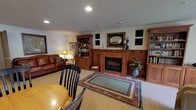 This single-floor space offers a well-appointed and comfortable layout designed for both relaxation and gatherings. The living area serves as the heart of the space, anchored by a classic wood-trimmed fireplace flanked by rich oak built-in bookcases and display cabinetry. Carpeted flooring, recessed lighting, and a textured ceiling contribute to a warm, inviting ambiance, while a patterned area rug adds a decorative touch. Seating includes a full leather sofa, a reclining armchair, and additional lounge furniture arranged for easy conversation and entertainment. A flat-screen television is neatly integrated into the built-in cabinetry, and a window with a roller shade provides filtered natural light and outdoor views. A dedicated dining area features a large rectangular table surrounded by upholstered chairs, positioned beside expansive sliding glass doors that overlook a green outdoor setting, flooding the room with natural light. The kitchen is fully equipped with oak cabinetry, a side-by-side refrigerator, a range, and a built-in microwave, complemented by a bar counter with seating. Throughout the space, warm neutral tones, wood accents, and carefully arranged furnishings create a cohesive, homey atmosphere suited for comfortable everyday living.