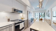 Unit 410 at 25 Gibbs Road offers a thoughtfully designed one-bedroom, one-bathroom residence spread across a single floor. The open-concept layout efficiently connects the kitchen, living, and dining areas, creating a cohesive and functional living environment bathed in natural light.

The kitchen is a standout feature, showcasing a central island with a white quartz countertop and bar seating, complemented by two-tone cabinetry in white and light wood. Stainless steel appliances — including a range, dishwasher, microwave, and refrigerator — are paired with a sleek tile backsplash and pendant lighting overhead.

The living room is bright and inviting, anchored by a large floor-to-ceiling window that frames urban views of the surrounding cityscape. Light hardwood flooring flows throughout, and the space is styled with a dark ribbed credenza, a round glass coffee table, and neutral-toned furnishings that enhance the airy atmosphere.

The bedroom continues the light aesthetic, featuring a large window with city views, mirrored sliding closet doors, and warm natural light that fills the room throughout the day.

The bathroom is finished with large-format beige tiles, a soaker tub with shower, and a sleek floating white vanity with under-mount sink, delivering a clean, spa-like atmosphere.

Overall, the residence presents a modern, minimalist design with a warm, neutral palette throughout.