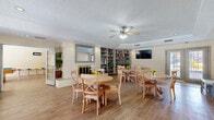 This single-floor community space is thoughtfully designed to encourage social interaction and relaxation. The main gathering area features warm wood-look flooring, painted brick accent walls, and a tray ceiling with recessed lighting and ceiling fans that lend the room an open, airy feel. Multiple round dining tables with cross-back chairs are arranged throughout, creating a welcoming atmosphere for group gatherings. A classic fireplace serves as a focal point along one wall, framed by a decorative mirror, while built-in bookshelves stocked with a curated collection of books wrap around an adjacent wall, adding a library-like charm.

A wall-mounted flat-screen television provides entertainment, and French doors along the exterior walls invite natural light and outdoor views into the space. The game room area extends the footprint of the space and accommodates a full-size pool table alongside a high-top dining table and bar-height chairs, perfect for casual recreation. Cozy reading nooks with tufted armchairs and pouf ottomans are tucked throughout, offering quieter spaces for relaxation.

A compact kitchenette supports the space with cabinetry, a sink, refrigerator, and countertop appliances. The overall ambiance is warm, inviting, and well-suited for community activities and everyday gathering.