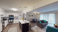 This single-floor residence is designed with a practical, modern aesthetic and an open-concept layout that seamlessly connects the kitchen, dining, and living areas. The kitchen is a standout feature, showcasing white shaker-style cabinetry, stainless steel appliances including a refrigerator, range, microwave, and dishwasher, along with a mosaic tile backsplash and sleek countertops. A central island with pendant lighting provides additional prep space and casual seating. Wood-style flooring flows throughout the main living areas, complementing the neutral gray walls.

The living room offers generous space, anchored by a ceiling fan and large vertical-blind-covered windows and a sliding glass door that invite natural light and outdoor views. A stacked washer and dryer unit is conveniently integrated within the living space.

Both bedrooms are carpeted for comfort, each featuring closet storage and windows with vertical blinds. The primary bathroom includes a vanity with a stone-look countertop, a framed mirror, and a glass-enclosed shower. The secondary bathroom offers a tub-shower combination with a dark vanity. The overall ambiance is clean, contemporary, and functional throughout.