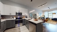 This thoughtfully designed single-floor residence combines modern aesthetics with functional living. The open-concept kitchen is a standout feature, showcasing two-tone cabinetry with crisp white upper cabinets and slate gray lower cabinets, accented by matte black hardware. Stainless steel appliances, including a range, microwave, refrigerator, and dishwasher, complement the quartz countertops. A peninsula with bar seating and a sleek linear pendant light serves as both a prep space and casual dining area, while a matte black faucet adds a refined touch to the undermount sink. Wood-look plank flooring flows seamlessly from the kitchen into the open living area, which is bathed in natural light through large windows with sheer curtains. The living room offers a welcoming ambiance with generous wall space ideal for artwork display. The bedroom continues the natural light theme with oversized windows and plush carpeting, creating a calm retreat. A dedicated office nook features bold accent wallpaper, floating wood shelves, and carpet underfoot, offering a creative and functional workspace. The bathroom is finished with tile surround, a tub-shower combination, and clean white cabinetry, maintaining the residence's consistent modern design language throughout.