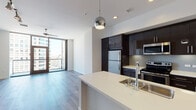 Located in the heart of Downtown Los Angeles, this modern residence at 801 S Hope St offers a thoughtfully designed open-concept layout finished with light hardwood flooring throughout. The kitchen serves as a central hub of the space, showcasing dark espresso cabinetry that extends to the ceiling for ample storage, stainless steel appliances including a refrigerator, range, and microwave, a subway tile backsplash, and a generous white quartz island with a built-in sink — illuminated by contemporary globe pendant lights. The open living area is bathed in natural light from expansive floor-to-ceiling windows and a glass balcony door that frames striking views of the Downtown Los Angeles skyline. A ceiling fan and track lighting add both functionality and style to the living space. The bathroom is clean and modern, featuring a dark-cabineted vanity with a white quartz countertop, a large mirror, and a tiled soaking tub and shower combination with neutral grey tile. White walls and crisp finishes throughout lend the space a bright, airy ambiance well-suited to contemporary urban living.