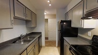 This single-floor space is organized into distinct living and functional areas, each with its own practical features. The living room is a spacious, open area finished with wall-to-wall carpeting in a neutral tone. A large sliding glass door fills the room with natural light and opens to a private balcony with an outdoor view of surrounding trees, offering a pleasant connection to the outdoors. A wall-mounted air conditioning unit and a baseboard heater along the perimeter ensure year-round climate control. The walls are painted in a soft, neutral gray, contributing to a calm and understated ambiance.

The kitchen follows a galley-style layout, efficiently designed with cabinetry lining both sides. Gray-toned cabinets with simple hardware provide generous storage space, complemented by dark laminate countertops and a double stainless steel sink. Black appliances, including a refrigerator and range, add a consistent visual contrast throughout the kitchen. Light tile flooring and track lighting overhead keep the space bright and functional. A passageway at the end of the kitchen connects to an adjacent carpeted area with a closet, adding additional utility to the overall layout.