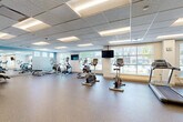 Located at 6000 NW 62nd Ave in Johnston, Iowa, this expansive single-floor amenity space spans a generously sized footprint designed to serve a wide range of resident needs. The centerpiece is a large, open exercise room outfitted with Matrix-brand cardio equipment, including treadmills, ellipticals, and stationary bikes, all arranged across durable rubber flooring. Large windows allow natural light to fill the workout areas while offering exterior views, and wall-mounted flat-screen TVs provide entertainment throughout. A dedicated yoga and stretching studio features light wood flooring, full-length mirrors, built-in shelving, and an inspirational wall display, with direct access to locker facilities through frosted glass doors. Adjacent to the fitness areas is a game room with a billiard table, comfortable seating, and floor-to-ceiling dark wood cabinetry for storage. A vending alcove provides convenient access to beverages and snacks. The space also includes a fully equipped pet grooming station with a stainless steel wash tub and professional-grade dryer. Connecting hallways feature tile flooring, stairwell access, and double entry doors, tying the entire amenity floor together with a clean, modern aesthetic accented by neutral tones and bold color highlights.