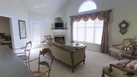 This two-story home showcases a beautifully appointed living room as its centerpiece. The space is defined by soaring vaulted ceilings that draw the eye upward to a striking arched transom window, which floods the room with natural light and adds an elegant architectural focal point. Large windows dressed with layered valances and drapes further enhance the brightness of the room while providing a refined, classic aesthetic.

The living room features a traditional fireplace framed by a decorative mantel, above which a television is mounted, seamlessly blending functionality with style. Ornate carved wooden furniture, including a generously sized sofa and accent chairs upholstered in neutral tones, anchors the room and contributes to its formal yet inviting character. Plush wall-to-wall carpeting adds warmth underfoot throughout the space.

A side door provides convenient access to an exterior area, allowing natural light to filter into the room from multiple directions. The overall ambiance of the home is one of traditional elegance, with carefully considered details — from the decorative drapery to the carved furniture — that speak to a classic and timeless design sensibility across both floors.