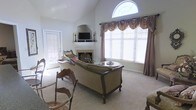 This two-story home showcases a beautifully appointed living room as its centerpiece. The space is defined by soaring vaulted ceilings that draw the eye upward to a striking arched transom window, which floods the room with natural light and adds an elegant architectural focal point. Large windows dressed with layered valances and drapes further enhance the brightness of the room while providing a refined, classic aesthetic.

The living room features a traditional fireplace framed by a decorative mantel, above which a television is mounted, seamlessly blending functionality with style. Ornate carved wooden furniture, including a generously sized sofa and accent chairs upholstered in neutral tones, anchors the room and contributes to its formal yet inviting character. Plush wall-to-wall carpeting adds warmth underfoot throughout the space.

A side door provides convenient access to an exterior area, allowing natural light to filter into the room from multiple directions. The overall ambiance of the home is one of traditional elegance, with carefully considered details — from the decorative drapery to the carved furniture — that speak to a classic and timeless design sensibility across both floors.