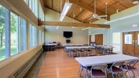 This single-floor community or event hall offers a welcoming and functional space well-suited for meetings, gatherings, and multipurpose use. The room's most striking architectural feature is its soaring vaulted ceiling, finished in warm natural wood with exposed timber beams that lend a rustic yet refined character to the interior. Ceiling fans are suspended from the rafters, ensuring comfortable airflow throughout the space.

Large windows line one wall, flooding the interior with abundant natural light and creating a bright, open atmosphere. The flooring is a warm hardwood, adding to the inviting feel of the room. Baseboard heating runs along the windowed wall, providing comfort across seasons.

The hall is set up with multiple rectangular folding tables and chairs, arranged to accommodate a variety of event configurations. A wall-mounted flat-screen television serves as a focal point for presentations or media display. A set of solid wood doors provides a formal entry point, complementing the overall natural material palette of the space.

Overhead fluorescent lighting supplements the natural light, ensuring the hall remains well-lit at all hours. The combination of high ceilings, open floor plan, and flexible furnishings makes this a practical and appealing venue for a wide range of community or organizational activities.