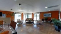 This single-floor space offers an open and inviting layout centered around a well-appointed lounge area. The interior is defined by warm orange accent walls that contrast with neutral furnishings, creating a vibrant yet comfortable atmosphere. Polished hardwood flooring runs throughout, reflecting the ample natural light that streams in through multiple large sliding glass doors and wide windows lining the perimeter of the room.

The seating area is thoughtfully arranged with a sectional sofa and coordinating armchairs positioned around a rectangular coffee table atop a decorative area rug. Potted plants and floral centerpieces add organic touches to the space. A framed piece of artwork adorns one of the orange accent walls, contributing to the finished, curated feel of the interior.

The textured ceiling features recessed lighting supplemented by HVAC vents, ensuring both comfort and functionality. The expansive windows provide panoramic views of what appears to be a waterfront setting, seamlessly connecting the interior to the outdoors. The overall ambiance is bright, open, and welcoming, suitable for communal gathering or relaxation.