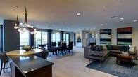 This elegantly designed single-floor residence showcases a seamless open-concept layout that effortlessly connects the living, dining, and kitchen areas into one cohesive and spacious environment. The interior is finished with wide-plank light hardwood flooring that runs throughout, lending warmth and continuity to the space. Recessed lighting is thoughtfully distributed across smooth ceilings, complemented by a striking contemporary chandelier that adds a sculptural focal point to the dining area. The dining space is anchored by a dark rectangular table surrounded by upholstered chairs, set atop a layered area rug that defines the zone with sophistication. Floor-to-ceiling sliding glass doors and expansive windows flood the interior with natural light while framing views of an outdoor terrace and the surrounding landscape beyond. The living area features a modern sectional sofa adorned with decorative pillows in deep teal and neutral tones, accompanied by sleek side tables and a curated arrangement of tall ceramic lamps and vases. A bold, colorful abstract artwork adorns the wall, adding a vibrant artistic accent. The kitchen, partially visible in the foreground, features dark cabinetry with clean contemporary lines. The overall ambiance is refined, modern, and inviting.