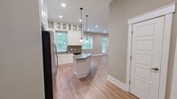 Located at 2324 McClintock Road in Charlotte's 28205 corridor, this well-appointed unit offers a cohesive open-concept layout that seamlessly connects the kitchen and living room. The kitchen is a standout feature, showcasing floor-to-ceiling white shaker cabinetry with glass-front upper panels, a mosaic tile backsplash, dark quartz countertops, a central island with pendant lighting, and a full suite of stainless steel appliances. Natural light filters in through multiple windows, lending the space a bright and airy feel throughout. Warm-toned hardwood-style flooring runs continuously through the main living areas, complementing the neutral taupe wall palette and crisp white trim. The living room is generously proportioned, with multiple windows and a glass-panel door providing views of the surrounding greenery. A set of sleek sliding barn-style doors with frosted glass panels leads to the bedroom area, adding a modern architectural touch. The bathroom is finished with tile flooring, a glass-enclosed shower, white vanity cabinetry, and dark countertops consistent with the overall design language. A walk-in closet with built-in wire shelving completes the unit, offering practical and organized storage solutions.
