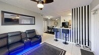 Located in Florida City, FL, this 2-bedroom, 3-bathroom single-floor residence offers a compact yet well-organized layout with a distinct modern design sensibility. The living room is a standout space, anchored by a large wall-mounted TV, a reclining leather sectional sofa with built-in blue LED accent lighting, and light tile flooring — creating a sleek, lounge-like atmosphere. Ceiling fans are present throughout, providing comfort suited to the Florida climate.

The open-concept kitchen and dining area features warm wood cabinetry, light countertops, and a peninsula with bar-height seating. Bold navy and white vertical striped walls define the kitchen pass-through, adding a graphic design element that energizes the space.

Both bedrooms are thoughtfully appointed with ceiling fans, closet storage, and direct access or proximity to bathrooms. One bedroom showcases a dramatic black tufted headboard and dark curtains for a moody, sophisticated feel, while the other maintains a cleaner, lighter palette with a wall-mounted TV. A third bathroom features an accessible roll-in shower with grab bars. A dedicated laundry area and additional utility space round out the practical offerings of this well-equipped home.