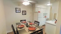 This single-floor space showcases an open-concept layout that seamlessly connects the dining area and kitchen, creating a cohesive and practical living environment. The dining area is anchored by a dark-finished table comfortably seating four, accented by a patterned area rug and set with terracotta-toned dishware, lending a warm and inviting atmosphere. A ceiling fan with an integrated light fixture overhead provides both ventilation and ambient lighting to the dining space. Framed wall art in neutral tones adds a decorative touch to the beige walls, contributing to the overall understated aesthetic. The adjacent kitchen features white cabinetry that extends to the upper walls, offering generous storage space. White appliances, including a refrigerator and stovetop, maintain a clean, cohesive look throughout the kitchen. A red kettle on the range introduces a pop of color. The flooring transitions between the spaces with light-toned wood-style flooring, enhancing the continuity of the open layout. Overall, the space presents a functional and neatly arranged interior with a casual, homey character.