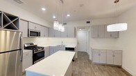 This single-floor space showcases a clean, contemporary design centered around a well-appointed kitchen. The kitchen is the standout feature, boasting an expansive white quartz island that serves as both a prep area and gathering spot. Sleek grey flat-front cabinetry lines the walls, providing ample storage, while stainless steel appliances — including a gas range, built-in microwave, and dishwasher — offer modern functionality. A double-basin sink sits beneath upper cabinets, complementing the bright white countertops and backsplash. Three stylish pendant lights hang above the island, adding a decorative focal point to the space. Recessed lighting throughout ensures the area is well-lit and welcoming. The flooring features light wood-look planks that run seamlessly through the space, adding warmth and continuity to the otherwise crisp, neutral palette. A hallway visible from the kitchen leads further into the unit, hinting at additional living areas beyond. The overall ambiance is fresh, bright, and move-in ready, with a cohesive modern aesthetic that balances practicality with contemporary style.