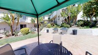 This inviting outdoor courtyard serves as a central communal gathering space nestled within a multi-story residential building complex. The patio is finished with stamped concrete tile flooring in a warm earth tone, providing a durable and attractive foundation throughout the space. Multiple stainless steel gas BBQ grills are stationed along a low retaining wall, offering residents ample cooking options for outdoor entertaining. A designated grill rules sign is posted nearby, reflecting the organized and community-minded nature of the space. Shaded seating areas are furnished with round dining tables and chairs beneath large green canopy umbrellas, ideal for outdoor meals. Additional lounge chairs are positioned across the patio, inviting relaxation in the sun. The courtyard is framed by mature tropical landscaping, including tall banana trees, palms, manicured hedges, and colorful drought-tolerant plants, all set against neatly maintained grass areas. The surrounding multi-story white stucco buildings with iron railings and exterior staircases create an enclosed, resort-like atmosphere. Trash receptacles are conveniently placed throughout the space, ensuring a clean and well-maintained environment. The abundant natural sunlight and lush greenery contribute to a bright, welcoming ambiance.