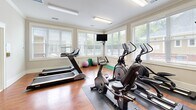 Located at 100 Avalon Park Cir in Asheville, NC, this fitness center offers a comprehensive workout facility within a efficiently utilized single-floor space. The room features warm hardwood flooring throughout, complemented by neutral-toned walls and crown molding that lend a polished, welcoming aesthetic.

The cardio area is positioned along a wall of large, blinds-fitted windows that allow generous natural light and views of the surrounding neighborhood, creating an open and airy atmosphere. This section houses multiple treadmills, elliptical trainers, and stationary bikes, along with a wall-mounted television for entertainment during workouts. Rubber floor mats are placed beneath equipment for added safety and stability.

The strength training area is equally well-appointed, featuring a multi-station cable machine, a seated press machine, an adjustable workout bench, and a tiered dumbbell rack stocked with a full range of free weights. Additional accessories, such as exercise balls, are also available.

Entry is provided through double glass-panel doors, reinforcing the bright and open feel of the space. Flush-mounted fluorescent lighting supplements natural light throughout. The overall design balances functionality with a clean, comfortable environment suitable for a full range of fitness activities.