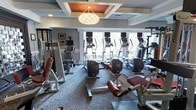 This fitness center is thoughtfully designed to offer a comprehensive workout environment within an elegant architectural setting. The space is anchored by a striking coffered ceiling with a warm, recessed orange accent panel and a decorative pendant light fixture, lending the room a refined, upscale ambiance that sets it apart from a typical gym.

Large windows line one wall, allowing abundant natural light to flood the interior and offering views of the surrounding greenery, creating an inviting atmosphere for exercise. The patterned carpet flooring adds a stylish, textured element underfoot throughout the space.

The cardio section is well-stocked with a row of elliptical trainers and additional cardio machines, all equipped with individual display screens. Strength training is equally well-represented, with a selection of resistance machines and weight benches featuring padded red upholstery positioned throughout the room.

Framed artwork on the walls and carefully considered finishes contribute to an environment that balances functionality with aesthetic appeal. The layout is open and organized, allowing for comfortable movement between equipment. Overall, this fitness center delivers a polished, fully equipped space suited for a wide range of exercise routines.