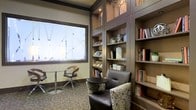 This single-floor reading lounge offers a thoughtfully designed space that balances functionality with comfort. The focal point is a floor-to-ceiling built-in bookshelf system finished in rich dark wood tones, featuring multiple open compartments stocked with books and decorative accents, including a sculptural tree motif integrated into the upper cabinetry. Subtle overhead lighting within the shelving units highlights the collection and adds warmth to the overall atmosphere.

A large frosted window with a geometric etched pattern floods the space with diffused natural light while maintaining privacy. Seating options are varied and inviting — a plush upholstered armchair with a patterned fabric sits alongside a small round side table, while a sleek bistro-style table with modern curved chairs offers an alternative spot for reading or quiet conversation. The neutral, earthy color palette of taupes, browns, and creams ties the furniture and built-ins together cohesively. Textured carpeting underfoot adds a layer of comfort and acoustic softness. Overall, the space conveys a quiet, refined ambiance well-suited for focused reading or relaxed solitude.