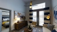 This single-floor space makes a strong visual impression through its notably high ceilings, which lend an airy, open quality to the interior. The living area is bathed in natural light, courtesy of large floor-to-ceiling windows dressed with bold navy and white striped curtains and slatted blinds, offering views of the surrounding exterior. A modern ceiling fan with a brass finish hangs prominently overhead, adding both function and a touch of contemporary elegance.

The living space is thoughtfully appointed with warm wood-tone flooring that grounds the room, complemented by a geometric-patterned area rug in blue and white tones. A substantial mid-century modern credenza in rich walnut anchors one wall, paired with an oversized abstract canvas artwork featuring vibrant blues and golds. Decorative accents, including a sculptural table lamp, ceramic vessels, and a potted succulent arrangement, add texture and personality to the space.

A cushioned accent chair positioned near the window creates an inviting reading nook, making the most of the natural light. The overall ambiance is modern and sophisticated, blending contemporary design elements with warm, earthy tones.