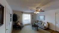 This single-floor space offers a warm and inviting atmosphere, centered around a well-appointed living room. The room features soft, neutral-toned carpeting that extends throughout, creating a cohesive and comfortable feel underfoot. A ceiling fan with an integrated light fixture serves as a functional focal point overhead, complemented by a textured ceiling that adds subtle visual character.

Natural light flows generously into the living room through a large window dressed with sheer white curtains, brightening the space and creating an open, airy ambiance. The layout comfortably accommodates a plush sofa adorned with decorative pillows, a patterned accent chair, and a classic dark-wood oval coffee table, all arranged to encourage relaxed conversation and everyday living.

Wall-mounted decorative accents and framed artwork contribute to the room's personalized character, while a lush indoor plant adds a touch of greenery. Neutral wall tones throughout keep the space feeling calm and versatile. The overall design strikes a balance between functionality and comfort, making it a welcoming environment suited to everyday living.