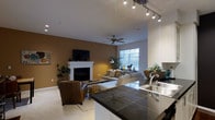 This single-floor residence offers an open-concept layout that seamlessly connects the kitchen and living areas, creating a spacious and cohesive environment. The kitchen is outfitted with dark tile countertops, a stainless steel sink, and crisp white cabinetry, complemented by track lighting overhead that provides focused, adjustable illumination. A raised breakfast bar defines the transition between the kitchen and the main living area, adding both function and visual separation.

The living room is anchored by a classic white-mantled fireplace set against a warm golden-brown accent wall, creating a welcoming focal point. A wall-mounted flat-screen television above the fireplace adds a modern touch. The space is furnished with a generous sectional sofa and a leather accent chair arranged on a neutral area rug, fostering a comfortable and inviting atmosphere. A ceiling fan with integrated lighting serves the center of the room, ensuring year-round comfort.

Natural light flows in through a large window, brightening the space and offering an airy feel. Potted greenery is thoughtfully placed throughout, adding organic texture and life to the interior. The light-toned carpeting throughout the living area contributes to the overall warmth and cohesion of this well-designed, functional home.