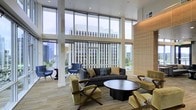 This single-floor lounge space is defined by its dramatic floor-to-ceiling glass walls, which flood the interior with abundant natural light and frame expansive views of the surrounding urban skyline. The double-height ceilings amplify the sense of openness, giving the space an airy, grand quality rarely found in communal settings.

The seating area is thoughtfully arranged around a sleek, dark circular coffee table, anchored by a charcoal sofa adorned with accent pillows in muted tones. Complementing the sofa are olive-green upholstered chairs with wooden X-frame bases, adding a mid-century modern touch, alongside a pair of navy blue accent chairs positioned closer to the windows—ideal for taking in the city views.

A warm wood-paneled accent wall provides textural contrast against the cool, glass-dominated perimeter, balancing the modern aesthetic with natural warmth. Light wood-toned flooring runs throughout, further enhancing the bright, welcoming atmosphere. Structural white columns punctuate the space, reinforcing its architectural character. Overall, the space conveys a polished, contemporary ambiance suited for relaxed gatherings or quiet reflection.
