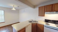 This two-floor residence offers a practical and comfortable layout spread across two levels connected by a carpeted staircase. The kitchen is well-appointed with warm oak cabinetry, light countertops, a double stainless steel sink, a dishwasher, a electric coil range with overhead ventilation, and a full-size refrigerator. A peninsula-style counter provides additional prep space and opens toward the adjacent living area, which features a combination of hardwood-style flooring and plush carpeting. A pendant light fixture adds a classic touch above this transitional space. The main living area is generously sized, with soft carpet, a ceiling fan with light fixture, and a large window dressed with vertical blinds that allows natural light to fill the room. The staircase leads to the upper level, where a spacious bedroom continues the carpeted flooring and includes its own ceiling fan and window with vertical blinds. The bathrooms feature oak vanity cabinetry, light countertops, and a mirrored vanity, with one bathroom offering a full tub-shower combination. Clean white walls and warm wood trim run consistently throughout the space, creating a cohesive and neutral aesthetic across both floors.