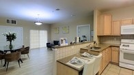 This single-floor residence offers a well-designed open-concept layout that seamlessly connects the kitchen, dining, and living areas into one cohesive living space. The kitchen is appointed with granite countertops, light wood cabinetry, and a tiled breakfast bar that doubles as a casual dining space, complete with seating and place settings. A stainless steel sink and ample counter space make the kitchen both functional and visually appealing. The breakfast bar transitions naturally into the dining area, creating a welcoming space for meals and entertaining. The living area beyond is bright and airy, benefiting from large sliding glass doors that allow generous natural light to fill the room and provide access to the outdoors. Light hardwood-style flooring runs throughout, adding warmth and continuity across the space. Neutral wall tones serve as a clean backdrop, accented by decorative wall art that adds a touch of personality. A pendant light fixture over the kitchen and a ceiling-mounted light in the living area provide well-distributed overhead lighting. The overall ambiance is calm, modern, and inviting, making this a comfortable and practical living environment.