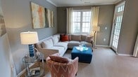 This single-floor space presents a well-appointed living area designed with a contemporary aesthetic and a warm, inviting atmosphere. The living room is finished with soft carpeting and neutral gray walls that serve as a versatile backdrop for colorful furnishings. Crown molding adds a refined architectural detail, lending the room a polished, traditional character.

A large, multi-pane window allows natural light to filter through sheer curtains, brightening the space and creating an airy feel. The room accommodates a generously sized sectional sofa in a light taupe tone, complemented by vibrant burnt orange and patterned throw pillows that introduce a lively contrast. A teal upholstered ottoman serves as a functional centerpiece, paired with a patterned accent chair in warm coral tones that adds visual interest.

Layered lighting is provided through a tall floor lamp and a table lamp on a glass-topped side table, enhancing the room's ambiance during evening hours. A large abstract canvas artwork adorns one wall, contributing a bold artistic focal point. Overall, the space balances comfort and style through a cohesive mix of textures, colors, and carefully chosen furnishings.